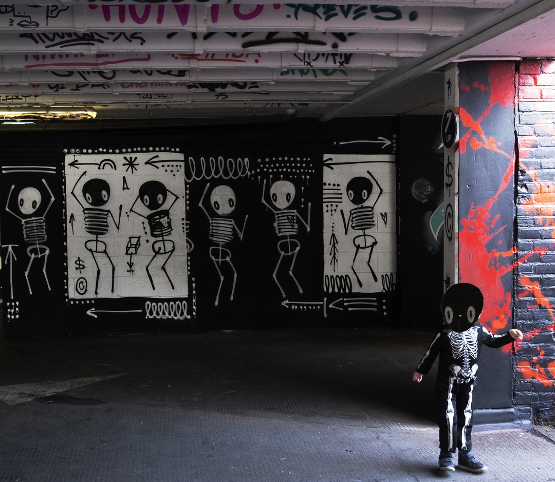 Skeleton paintings dance on a wall with young boy in skeleton costume in front, located in Penge multi story car park